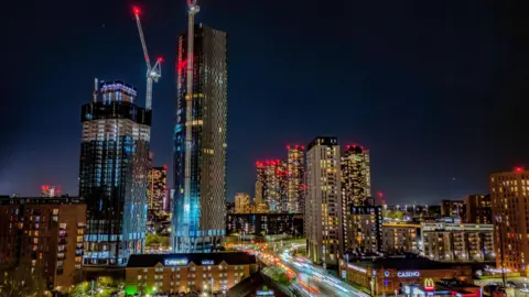 PA/Peter Byrne An at-night image of Manchester's skyline with several high-rise buildings lit up in yellow and red