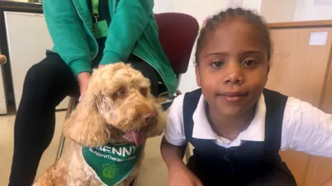 BBC Benny the dog has a green scarf around his neck and Dianne is wearing a black and white short-sleeved dress as part of her school uniform.