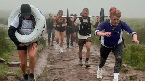 Sian Donoghue A group of two men and three women walking up a steep cobbled mountain path. One man is carrying a large rubber tyre, while another man is holding a large dumb bell.