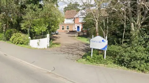 The outside of Darsdale Home for the Blind, showing a large building in the distance, a sign to the right, and lots of trees and foliage. There is a path down to the building.