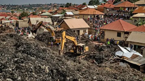 AFP Two yellow diggers are seen amidst the rubbish of Kiteezi with houses seen behind them and a crowd of onlookers - Kampala, Uganda, August 2024.