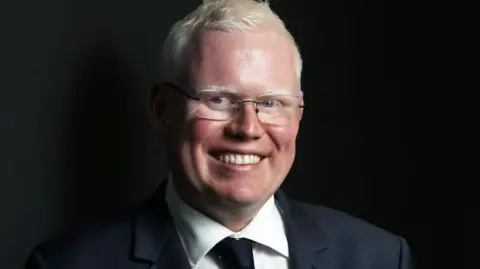 A man with white blond hair and glasses smiles at the camera