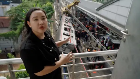 Liza stands above the train crash site and points to it as she speaks