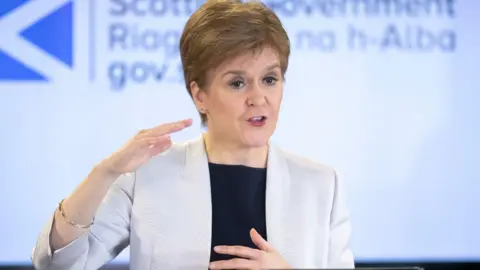 PA Media Nicola Sturgeon, who has short fair hair, speaks at a podium with her right arm raised. She is wearing a white jacket over a black top. 