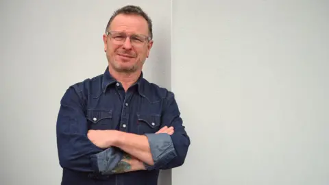 Mark looking at the camera with his arms folded. He is wearing a blue denim shirt and has his arms crossed, and has short cropped brown hair