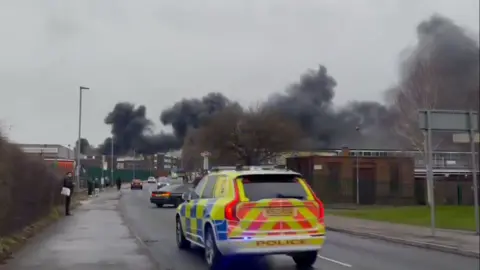 Marcus Reeves A police car drives to the scene of the factory fire with black smoke in the distance