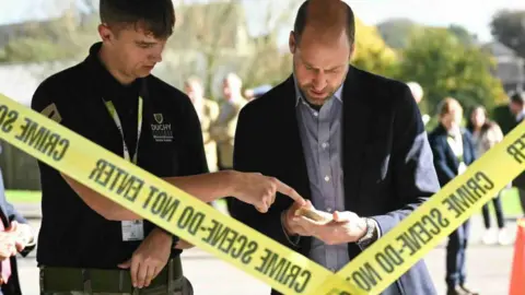 A young man points to an object in the hands of the Prince of Wales, who is wearing a dark jacket and blue shirt who is looking at the object with two strips of yellow tape in the foreground saying crime scene.