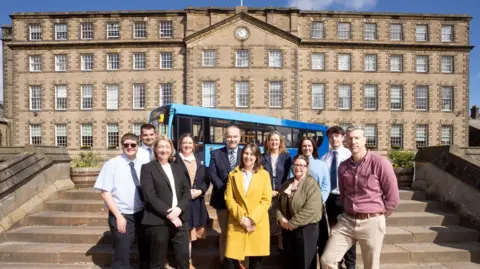 Councillor Karen Allison (centre) with representatives from the county’s tourist attractions, alongside officers from Durham County Council’s integrated passenger team and representatives from Northstar, the bus company which will be operating the services. They are standing on steps in front of the grand Ushaw historic house which is made of grey stones and lined with rows of square windows.
