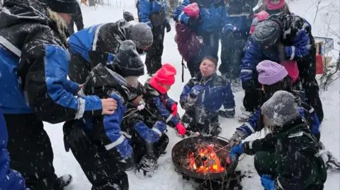 Paula Hudgell A group of adults and children sitting around a camp fire in the snow, roasting marshmallows. The children's faces are obscured so they cannot be identified.