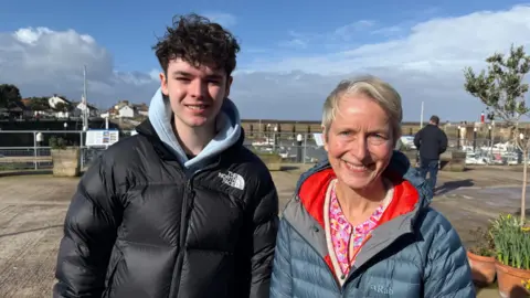 Younger chap on the left in a black North face puffer jacket coat. Woman stood next to him a blue coat and she is smiling at the camera.