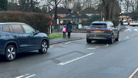 A road with cars on it. A mother and child walk on the pavement next to it, near a school.