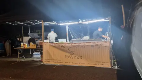 National Grid A welfare van offering hot food and drinks to customers. There are two people behind the catering stand and a white van on the right.