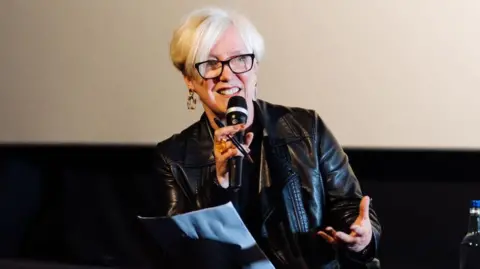 Getty Janice Forsyth in a leather jacket and wearing dark plastic glasses speaking at an event. She is holding a microphone and smiling, her hand waving in the air.