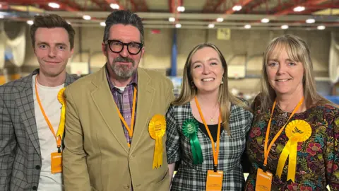 Four members of the Hunt family stand next to each other. From left to right is Nathan, Alan, Georgie, and Jacqueline. Nathan is wearing a white T-shirt and chequered blazer. He is also wearing a Liberal Democrat rosette. Alan is wearing a purple chequered shirt and a green coat, he is also wearing black framed glasses and a Liberal Democrat rosette. Georgie has long blonde hair and is wearing a black and white dress, a Green Party rosette and a lanyard. Jacqueline is wearing a floral dress and a Liberal Democrats rosette. She has long blonde hair and a fringe.