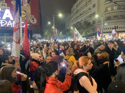 Male and female protesters hold Serbian flags and their mobile phones in the air.