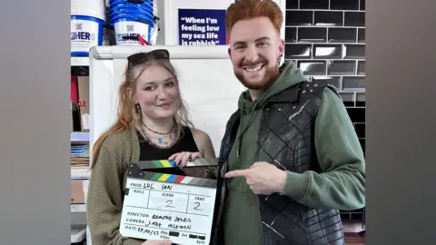 Phoebe Ashley A lecturer with a ginger quiff and beard smiles as they point at a clapperboard being held by a female university student with long blonde hair. The lecturer is wearing a black leather gilet over the top of a green hoodie, while the student is wearing a green cardigan over a black vest top and a stack of necklaces.
