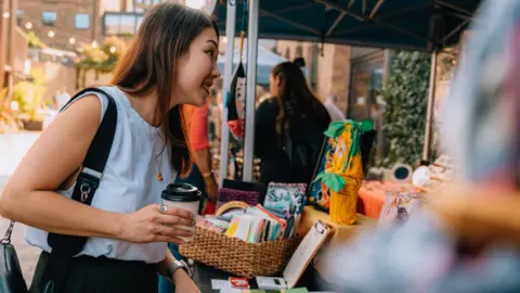 A woman with long brown hair is holding a coffee cup and is looking at a market stall with books for sale. 