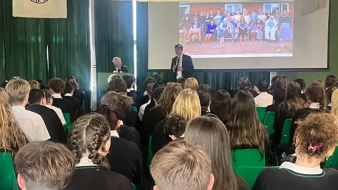 A group of school children listening to Steven Frank talk from a stage.