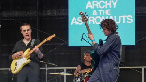 Three young male musicians perform on stage at Lincoln Live. Two guitarist are wearing navy blue boiler suits and the drummer is dressed in a black T-shirt. The band's name "Archy and the Astronauts" is on a light blue screen in the background.