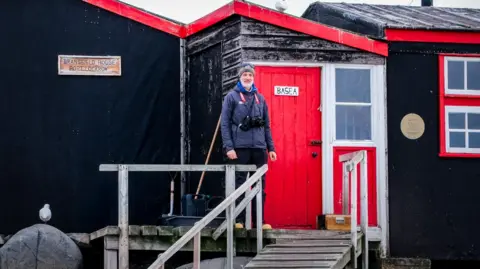 UKAHT/Pete Watson A man is standing in front of a black, snowy building wearing a coat and a hat. The door is painted red and says "base A" on it. There is a ramp leading away from the door.