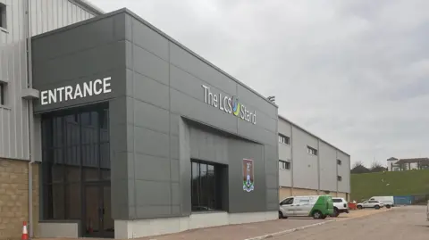 James Burridge/BBC The entrance to the new stand featuring Northampton Town's crest on the left hand side of the entrance. Above the glass doors, a sign reading "The LCS Stand" can be seen. 