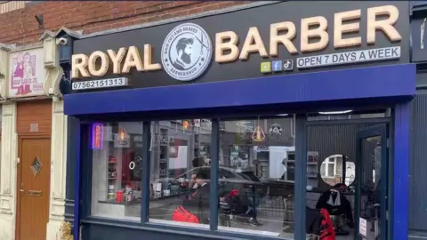 Shahram Hosseini front of barber shop - blue with gold letters. sign reads Royal Barber, open 7 days a week.