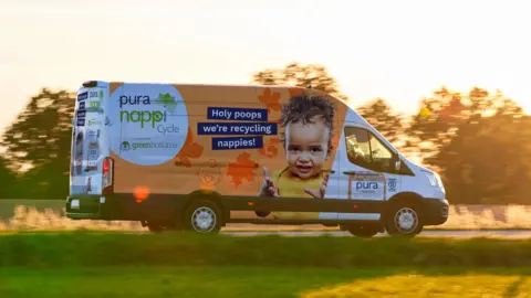 NappiCycle NappiCycle van seen driving by with trees in the background. The van is white with orange painting around the sides. On the side we're looking at, there's a picture of a baby with the text Holy poops we're recylcing nappies written in blue boxes in a white font.