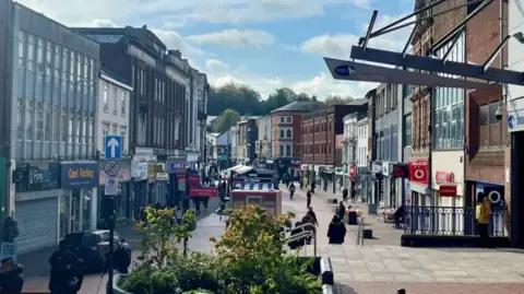 Walsall high street, with shops of different colours lining the pedestrianised route, which shows shoppers in the distance, with a flowerbed in the foreground and a car parked on the left. 