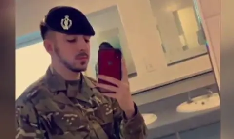 Selfie of Kyle Clifford wearing his army uniform and beret in a bathroom. He is holding a red phone and has a dark, close-cropped beard and moustache.