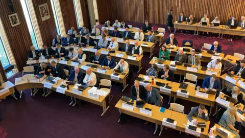 Laura Coffey/BBC A bird's eye view of about 60 councillors sitting at five rows of tables in a horseshoe shape in a council chamber. The carpet is a deep red and the walls are wooden panelled. There is a separate row of tables at the back of the room with half a dozen people sitting there.