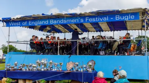 St Dennis Band A brass band plays on a stage during a competition called the West of England Bandsmen's Festival in Bugle. A banner with the name of the contest is above the stage. A table with dozens of trophies on it is in front of the stage.
