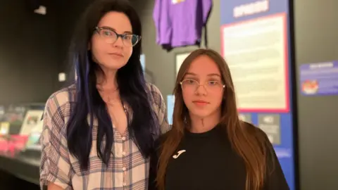 Jon Wright/BBC A woman with long dark hair and glasses stands next to her daughter, who also has long hair. They are looking at the camera. Behind are some artefacts on the wall as part of a museum exhibition.