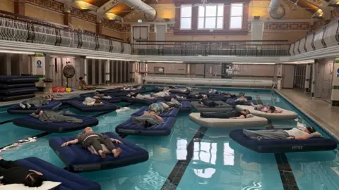 About a dozen clothed people lying on inflated mattresses, floating in an ornate indoor swimming pool. The water looks blue.
