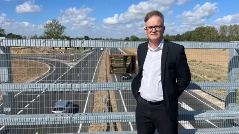 Qays Najm/BBC Chris Griffin wears a dark-coloured suit and a white shirt. He is standing on the new bridge. A car is passing underneath it on the dualled carriageway.