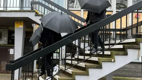 Three people walking down a set of steps holding umbrellas. They are all wearing black clothing. 