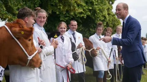 Prince William wearing a navy suit standing outside in a field at the Royal Norfolk Show pointing at a brown cow.  A group of people wearing white lab coats over shirts and ties line up next to the cow.