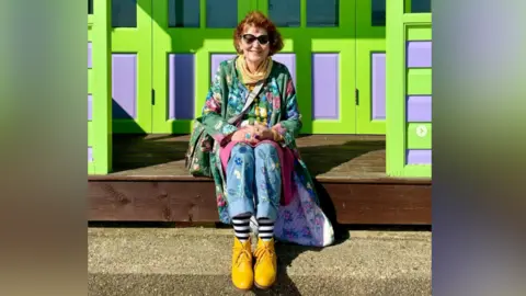 Contributed Marcia Riddington with short red hair and black sunglasses sits on a ledge outside a bright purple and green beach hut. She is wearing a green dress with flowers sewn into it and blue trousers with yellow boots.