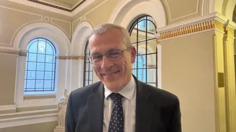 A man with short grey hair, wearing glasses and a suit, pictured from the shoulders up with windows and a statue behind him.