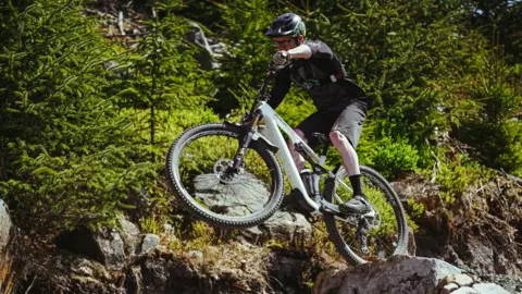 Forestry England Man wearing glasses and black helmet on a mountain bike in the air. The terrain is grassy and rocky. 