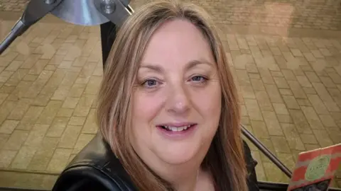 A woman with light brown shoulder length hair is wearing a black jacket and sitting down