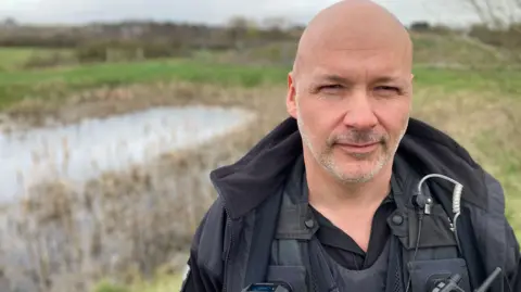 Det Con Aaron Flint standing next to the pond. He is wearing uniform and has short hair and a stubbly beard. The shot of the pond is out of focus.