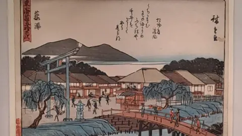 A traditional Japanese woodblock print depicting a scenic town near water. In the foreground, a curved wooden bridge spans a river, with several people walking across it. To the left, a large torii gate and a stone lantern stand near willow trees. 