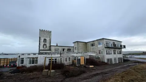 A derelict building, the former Castletown Golf Links Hotel, a large white building, there is mud in front, you can see the sea in the background. 
