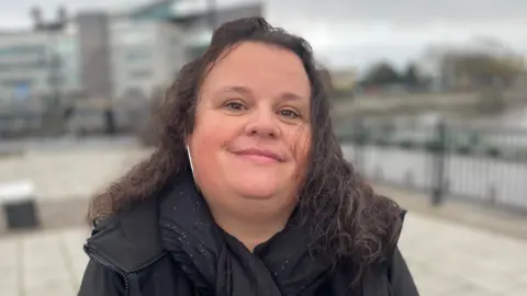 A woman with long dark hair wearing a black scarf with white specks and a black coat. It is a head and shoulders shot and she is stood outside, she is in focus while the outside backdrop is blurred.