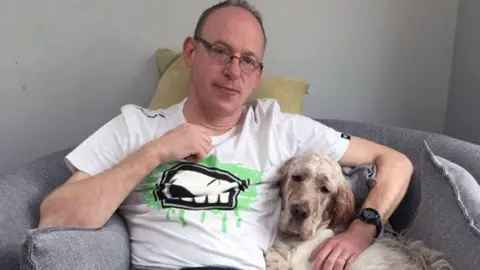 A man in glasses and a white t-shirt sits holding a dog
