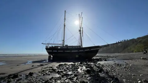 Julie Arthur A side-on view of a traditional tall ship sitting upright on a beach with the tide in, run aground. It is a gloriously sunny day with blue skies.