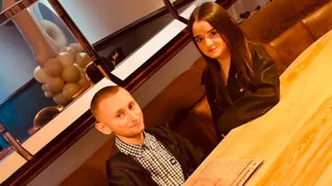 The pair are sat at a wooden table in a restaurant smiling at the camera. Lilly has long dark hair and Daniel has a shaved head. There is a mirror behind them.