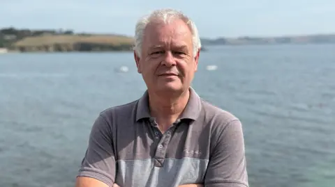 Nev has white hair and wears a grey polo short - he stands with the sea and coast behind him. 