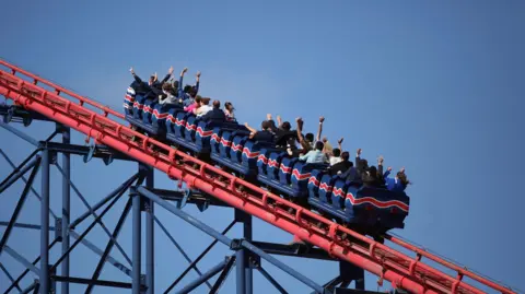 Getty Images A blue rollercoaster with a red stripe along the side makes it way up a red track with passengers on board holding their hands in the air.