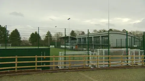 A large green pitch surrounded by green fencing. There is a small grandstand next to a large silver building overlooking the pitch